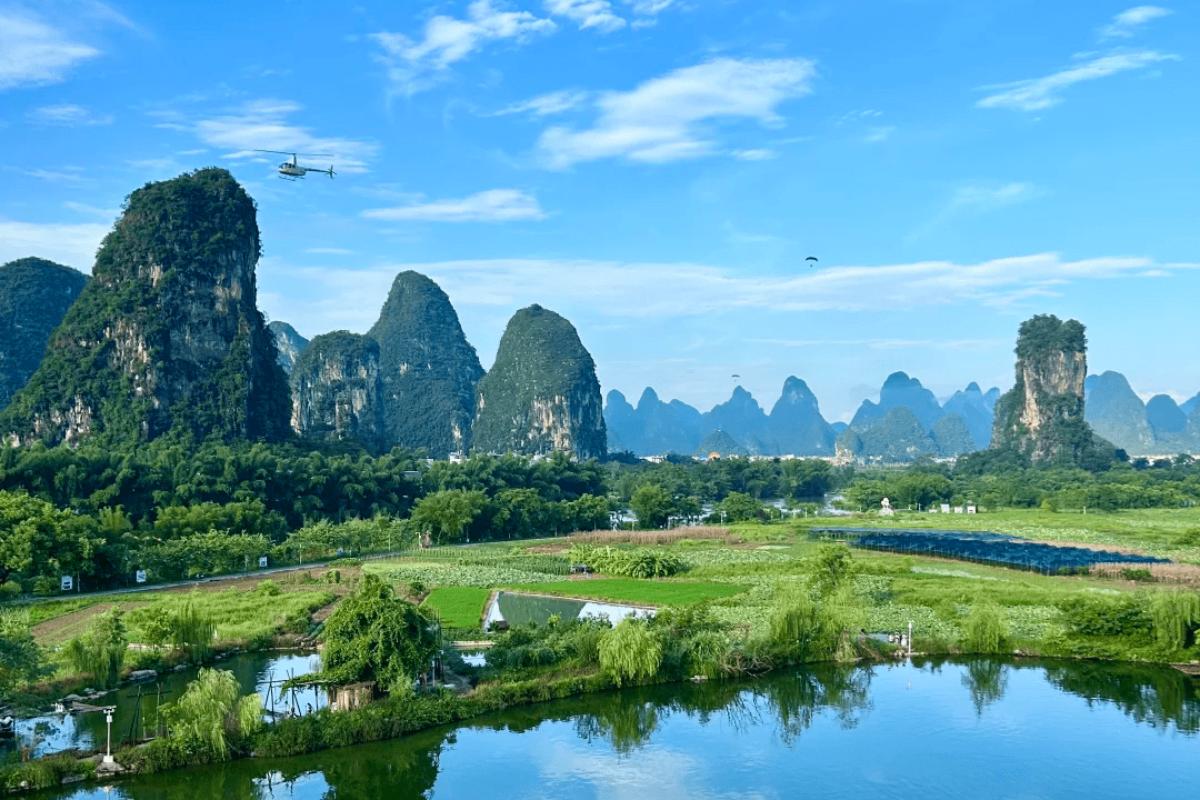 Scenic karst skyline beyond lush farmland and calm water, captured on a sunny day with aircraft visible overhead