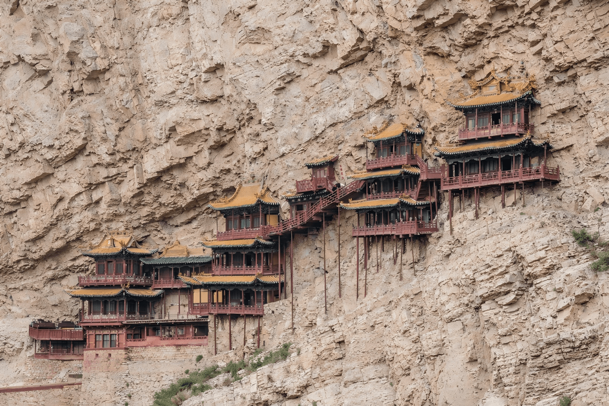 A historic multi-level wooden temple complex built into a steep rocky cliff, featuring traditional Chinese architecture with red pillars, golden roofs, and interconnected stairways suspended above the mountainside.