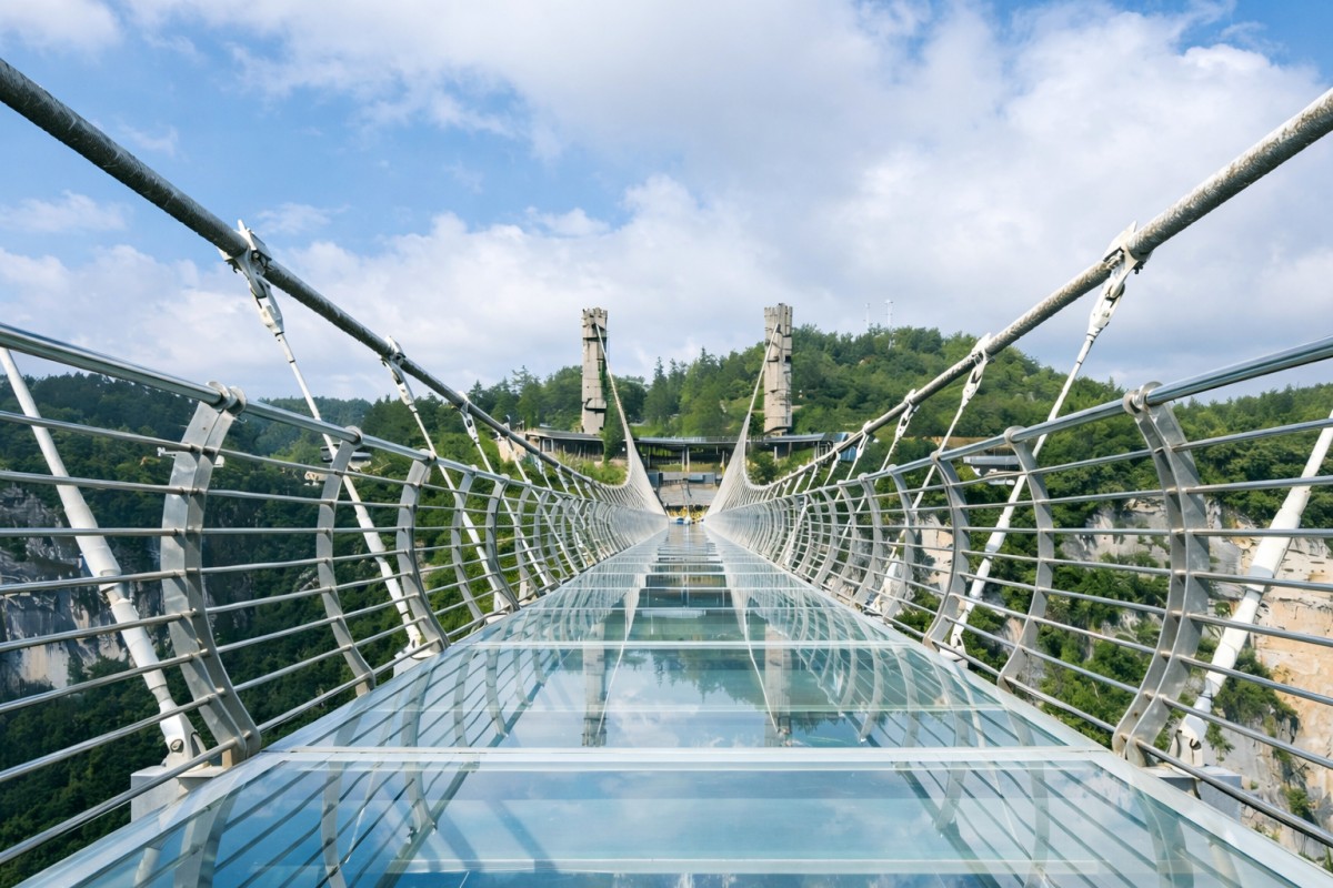 A modern glass-bottom suspension bridge stretching across a deep mountain gorge, with steel cables and railings leading toward forested hills under a partly cloudy blue sky.