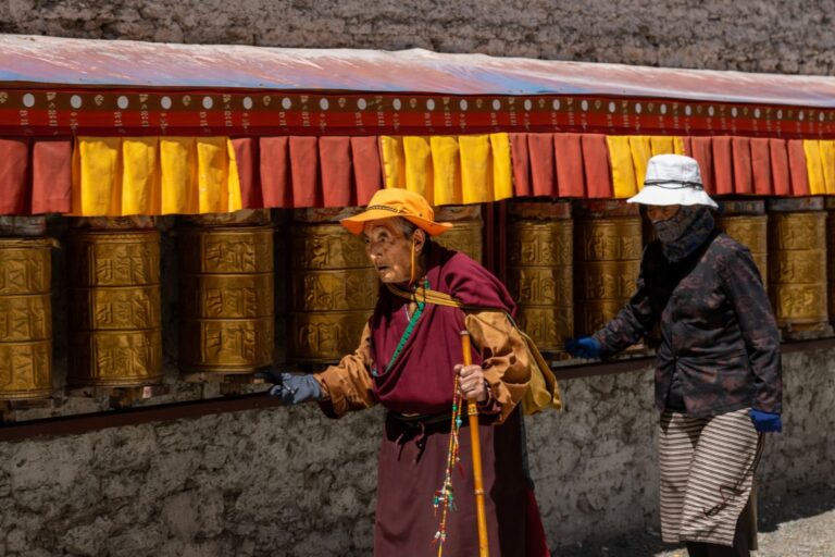 Two elderly women walking beside a row of golden Tibetan prayer wheels, spinning them as they pass, dressed in traditional clothing and hats under a red and yellow canopy.