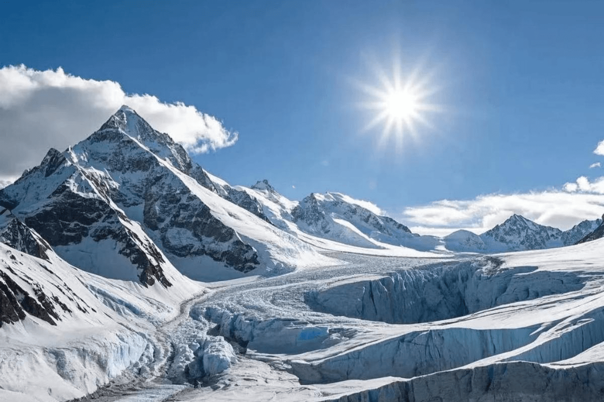 A vast glacier flows through a rugged mountain range covered in snow, with sharp peaks rising under a clear blue sky and a bright sun shining overhead.