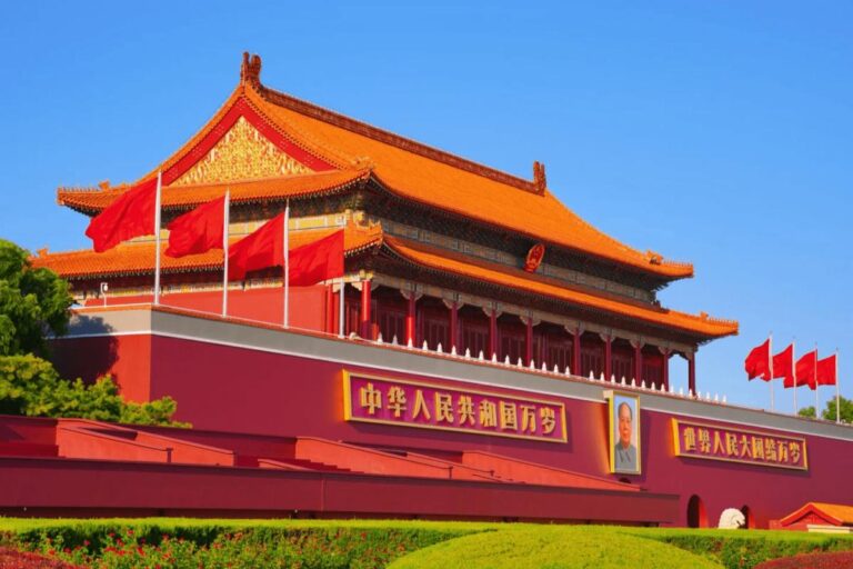 Tiananmen Gate with an orange-tiled roof, red flags flying, and a large portrait on the front wall above landscaped gardens.