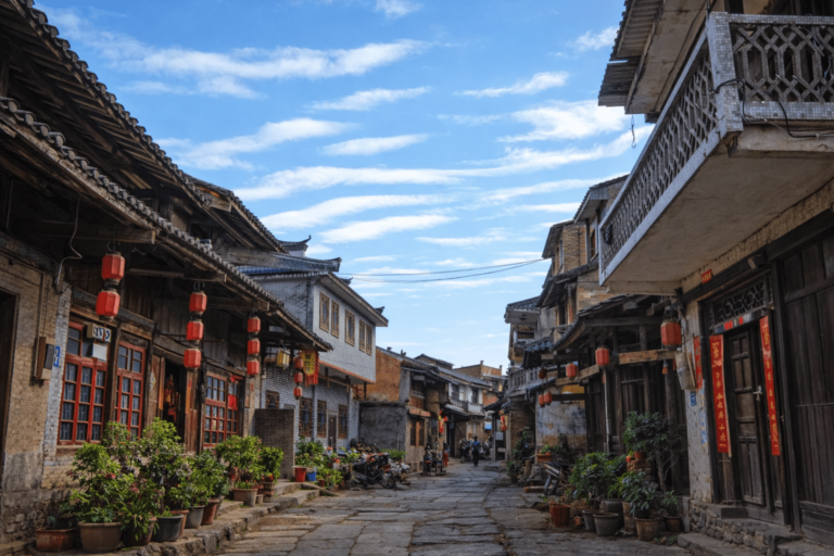 Stone-paved street lined with traditional wooden and brick buildings decorated with red lanterns and potted plants under a bright blue sky in a historic Chinese town.