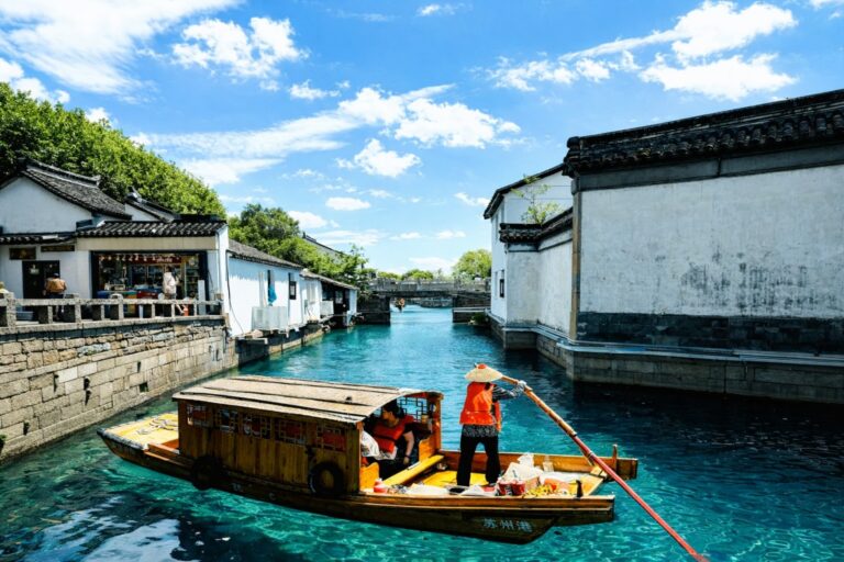 A wooden boat gliding through a narrow canal between whitewashed traditional houses in a historic Chinese water town under a blue sky.