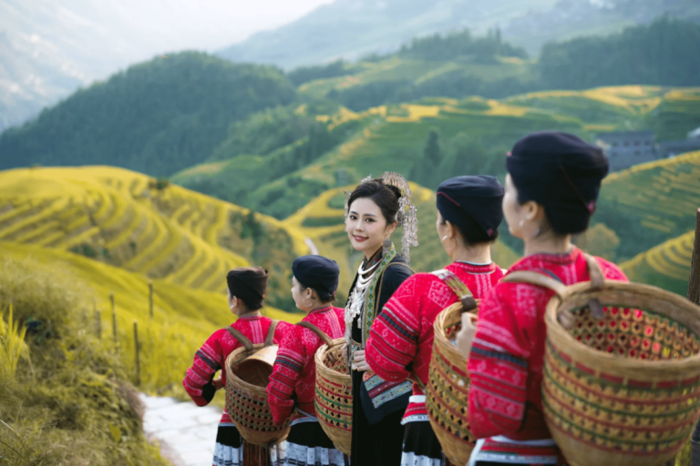Group of women wearing colorful traditional ethnic clothing and woven baskets standing on a path overlooking golden terraced rice fields and rolling green hills in a mountainous countryside.
