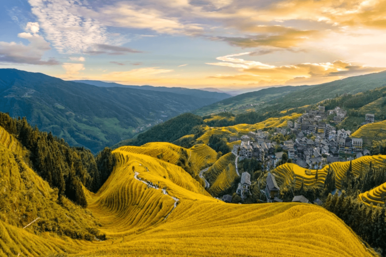 Sweeping view of the golden Longji rice terraces at sunset, with layered fields covering the hillsides and a traditional mountain village nestled among the terraces.