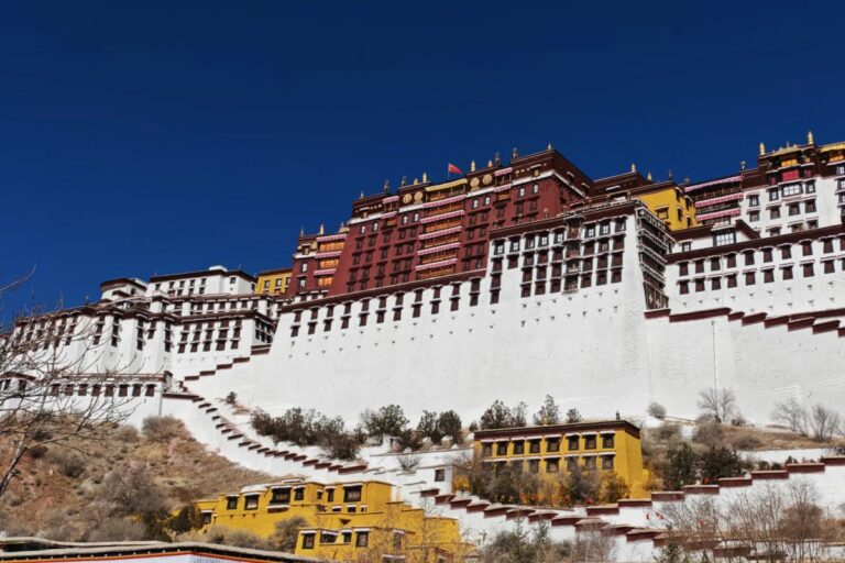 A grand palace complex with white walls and red and gold upper buildings rising on a hillside, set against a deep blue sky with smaller yellow structures at the base.