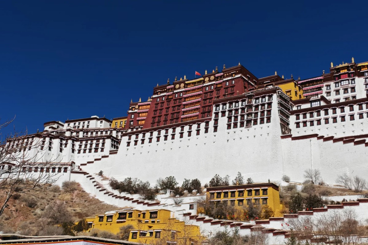 A grand palace complex with white walls and red and gold upper buildings rising on a hillside, set against a deep blue sky with smaller yellow structures at the base.