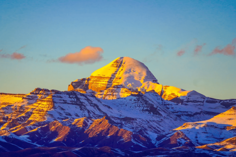 The towering Mount Kailash, illuminated by the warm glow of the setting sun. Its snow-covered peak contrasts against the rugged landscape below, with deep mountain slopes and a clear, colorful sky in the background. The iconic pyramid-shaped summit stands majestically amidst the serene Tibetan plateau.