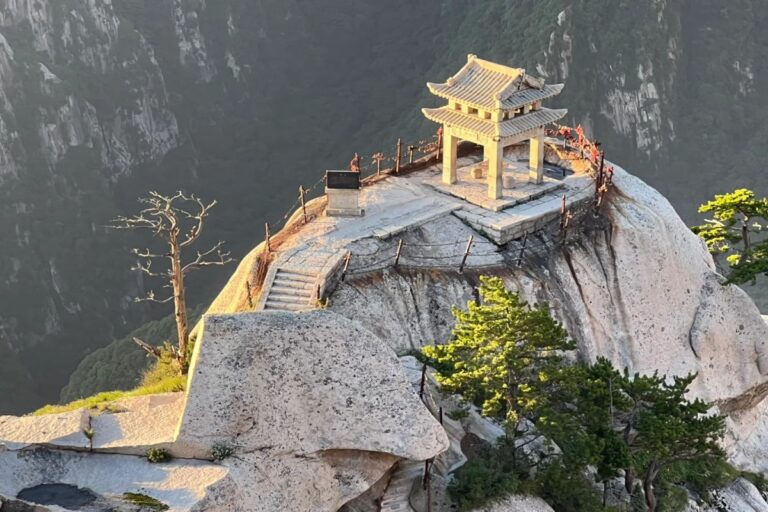 A small traditional Chinese pavilion perched dramatically on a sheer granite peak at Mount Hua (Huashan). Stone steps and iron chains cling to the cliff edge, while steep forested valleys fall away into mist below. Bathed in warm light, the isolated pavilion embodies both serenity and danger, capturing Huashan’s reputation as one of China’s most breathtaking and perilous sacred mountains.