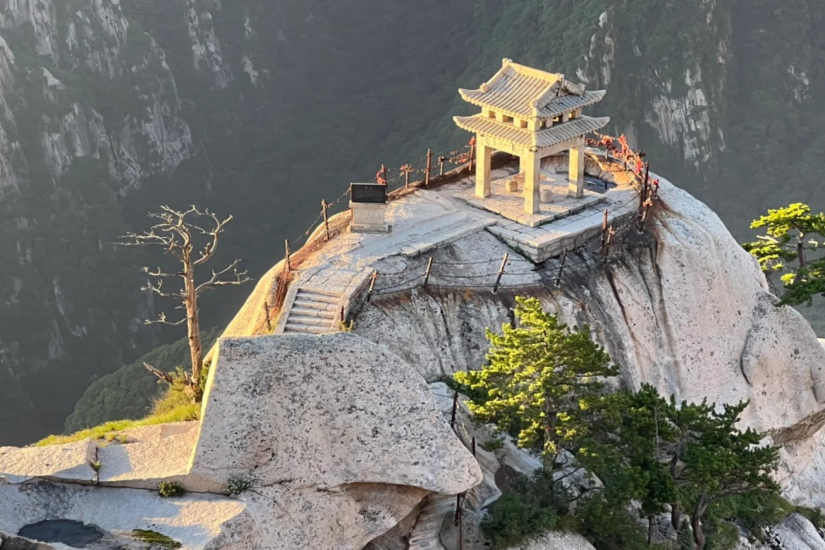 A small traditional Chinese pavilion perched dramatically on a sheer granite peak at Mount Hua (Huashan). Stone steps and iron chains cling to the cliff edge, while steep forested valleys fall away into mist below. Bathed in warm light, the isolated pavilion embodies both serenity and danger, capturing Huashan’s reputation as one of China’s most breathtaking and perilous sacred mountains.