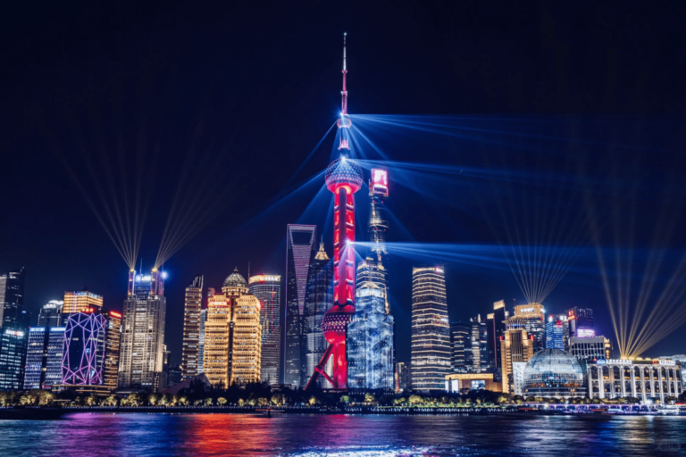 Night view of Shanghai’s skyline along the Huangpu River, featuring the illuminated Oriental Pearl Tower with colorful light beams and reflections on the water.