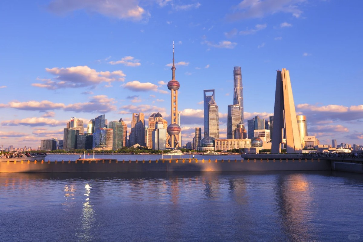 The Shanghai skyline at sunset, featuring the Oriental Pearl Tower and modern skyscrapers reflected in the calm waters of the Huangpu River under a blue sky with scattered clouds.