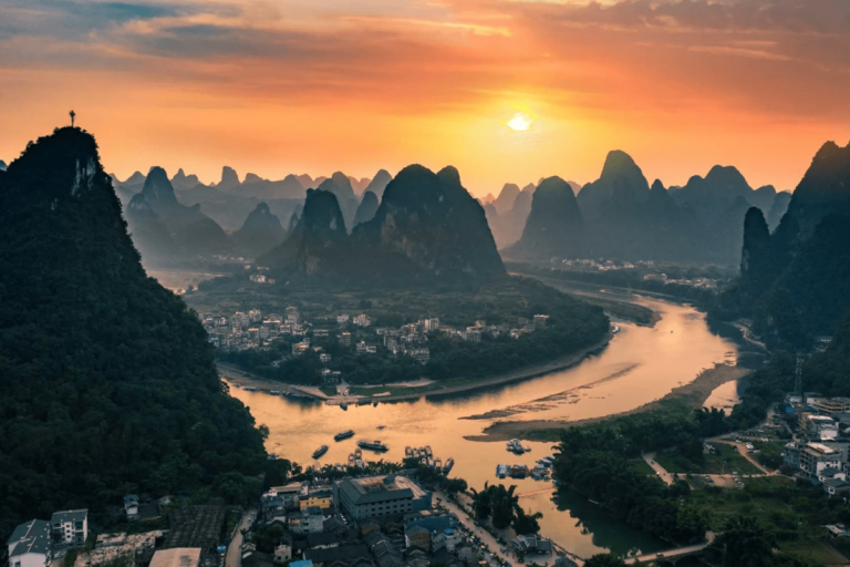 A panoramic view of a winding river flowing through a town surrounded by dramatic karst mountains at sunset. The golden sun casts warm light over the landscape, reflecting on the river and creating silhouettes of the layered peaks in the distance.
