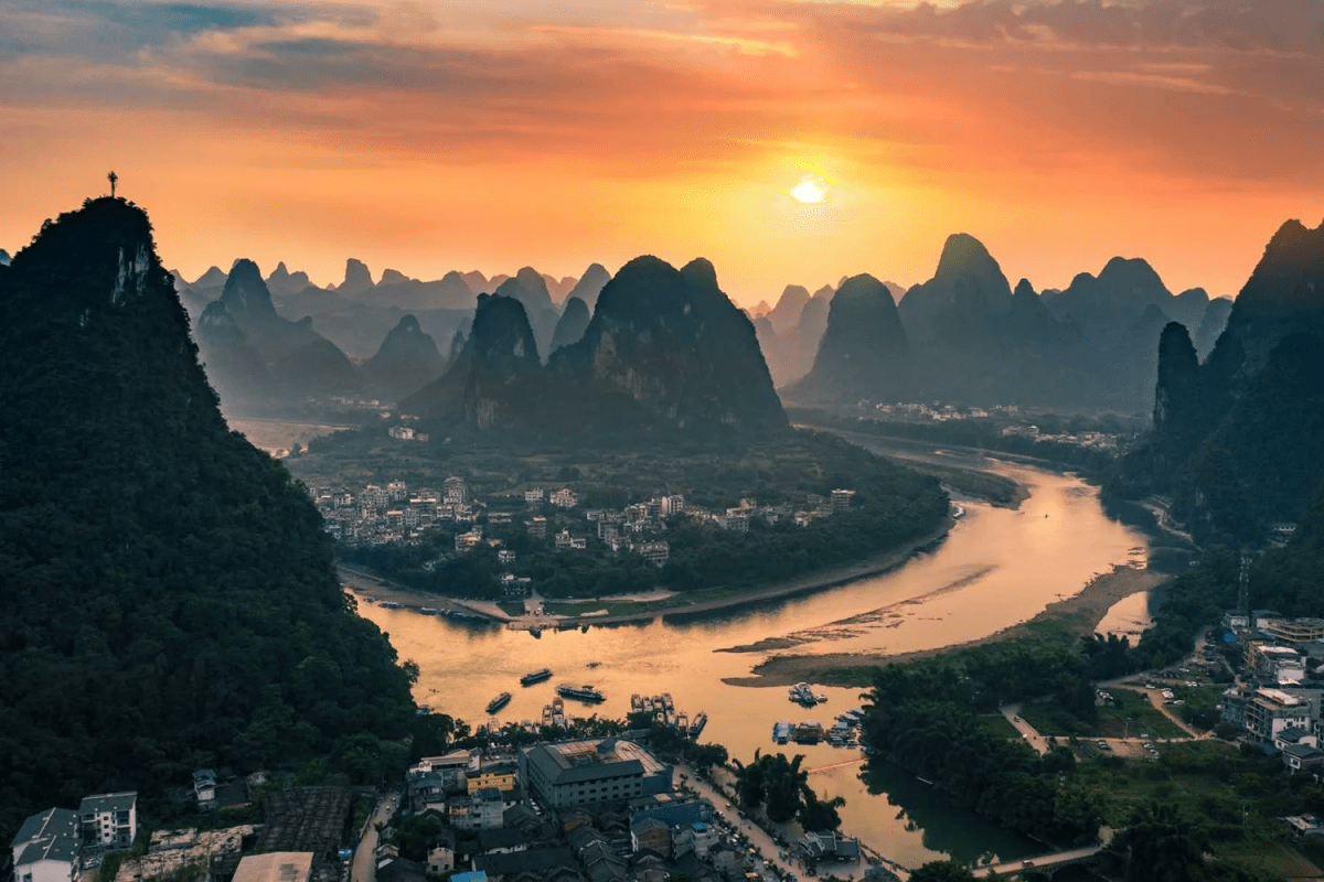A panoramic view of a winding river flowing through a town surrounded by dramatic karst mountains at sunset. The golden sun casts warm light over the landscape, reflecting on the river and creating silhouettes of the layered peaks in the distance.