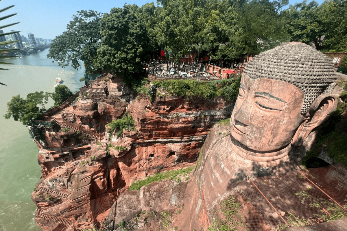 The Leshan Giant Buddha, carved into a cliff, gazes over the river. Surrounded by lush trees and a modern cityscape, the statue stands as a testament to ancient Buddhist art and history.