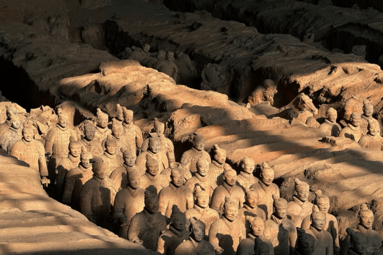 Rows of life-sized Terracotta Warriors stand partially excavated in an earthen pit, illuminated by warm light that highlights their detailed armor and facial features. The ancient clay soldiers are arranged in tight formation, revealing the scale and craftsmanship of the Terracotta Army, created over 2,000 years ago to guard China’s first emperor in the afterlife.