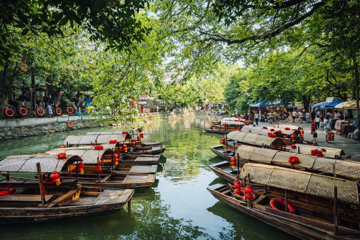 Wooden canal boats decorated with red lanterns are moored along a tree-lined waterway, with market stalls and visitors gathered on the riverbanks in a vibrant Chinese water town.
