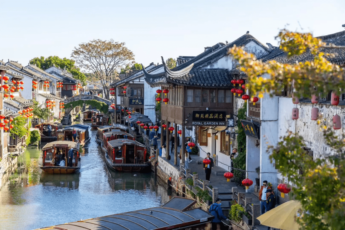A lively Chinese water town with traditional white-walled buildings and black-tiled roofs lining a narrow canal, red lanterns hanging along the streets, wooden boats cruising the water, and visitors walking beside the canal on a sunny day.