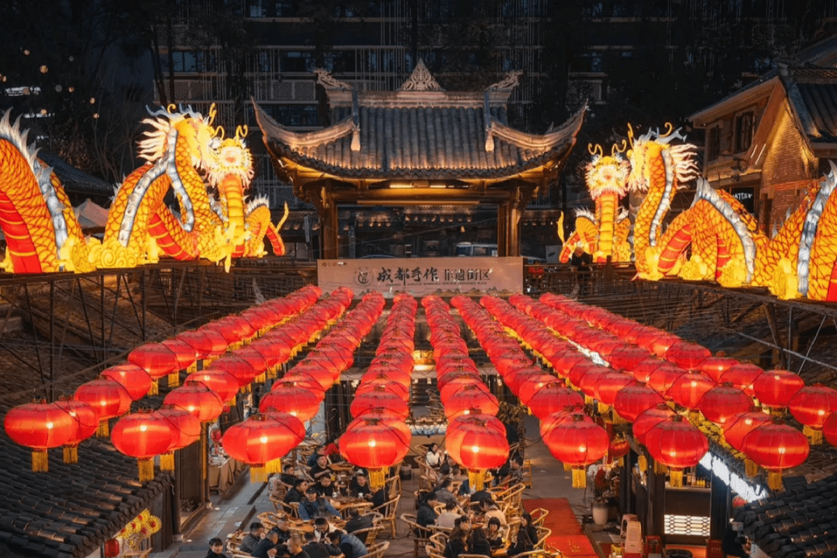 Rows of red lanterns hanging in a traditional courtyard at night, with illuminated dragon decorations and people below