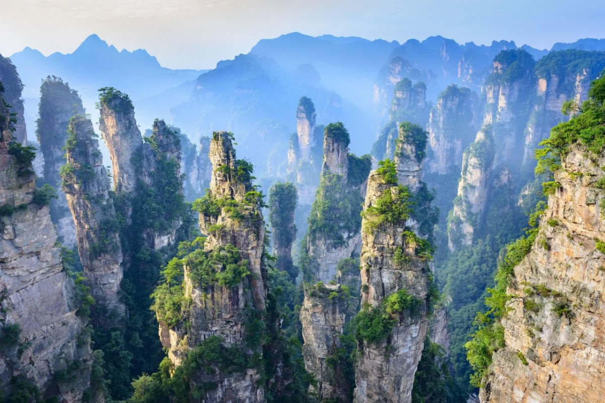 A breathtaking view of the Zhangjiajie Avatar Mountains in China, with towering, jagged peaks shrouded in mist. The lush greenery on top of each mountain adds a vibrant contrast to the rocky structures. The mountains appear to rise dramatically into the sky, creating a surreal landscape reminiscent of the floating mountains seen in the movie Avatar. The scene is peaceful and awe-inspiring, with layers of distant peaks and a hazy atmosphere.