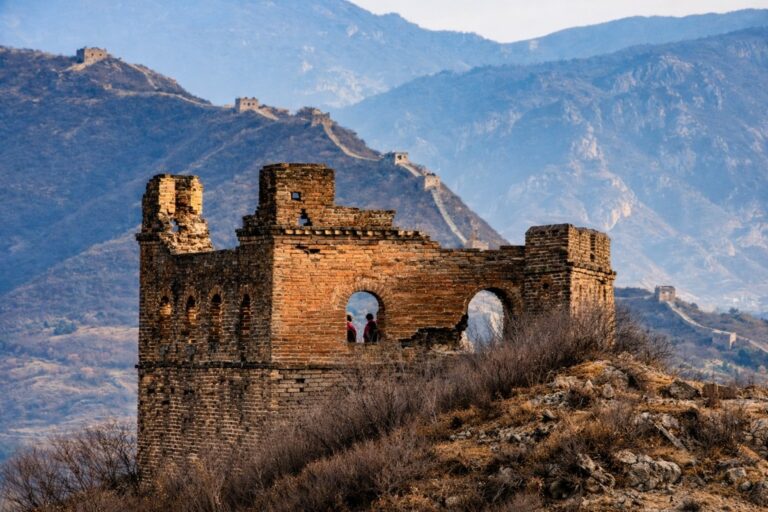 Ruined brick watchtower with two people inside, set against distant mountain ranges and the winding Great Wall in the background.