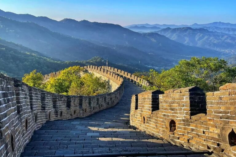 Winding stone pathway of the Great Wall with crenellated walls, surrounded by green trees and layered mountain ranges under a clear sky.
