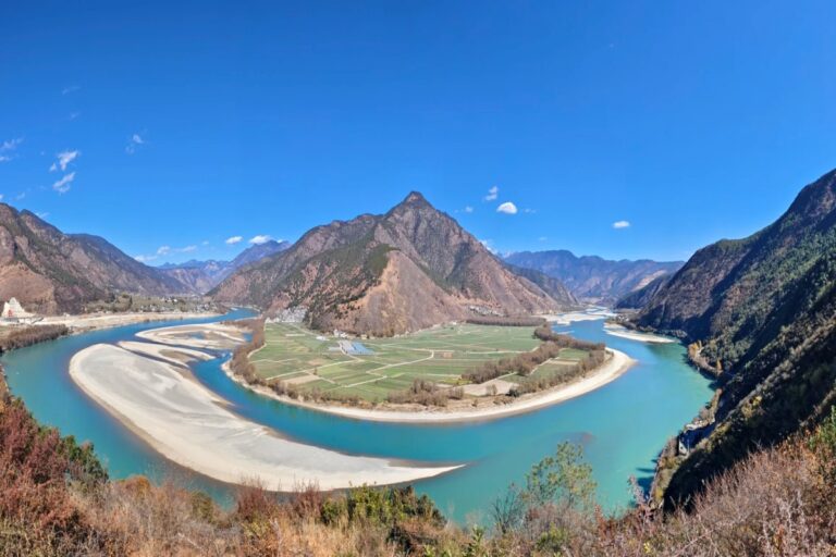 A turquoise river winding through a mountain valley with farmland on the inner bend and steep forested slopes on both sides under a clear blue sky.