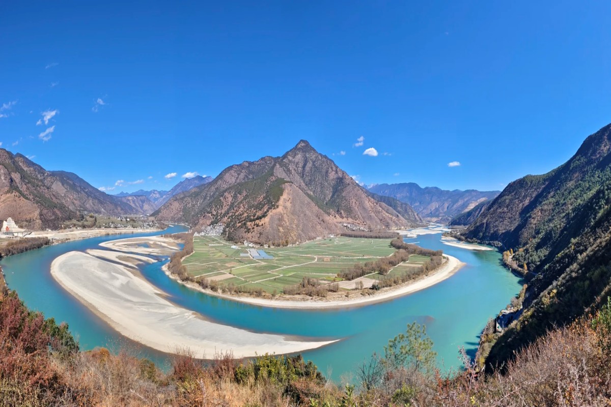 A turquoise river winding through a mountain valley with farmland on the inner bend and steep forested slopes on both sides under a clear blue sky.