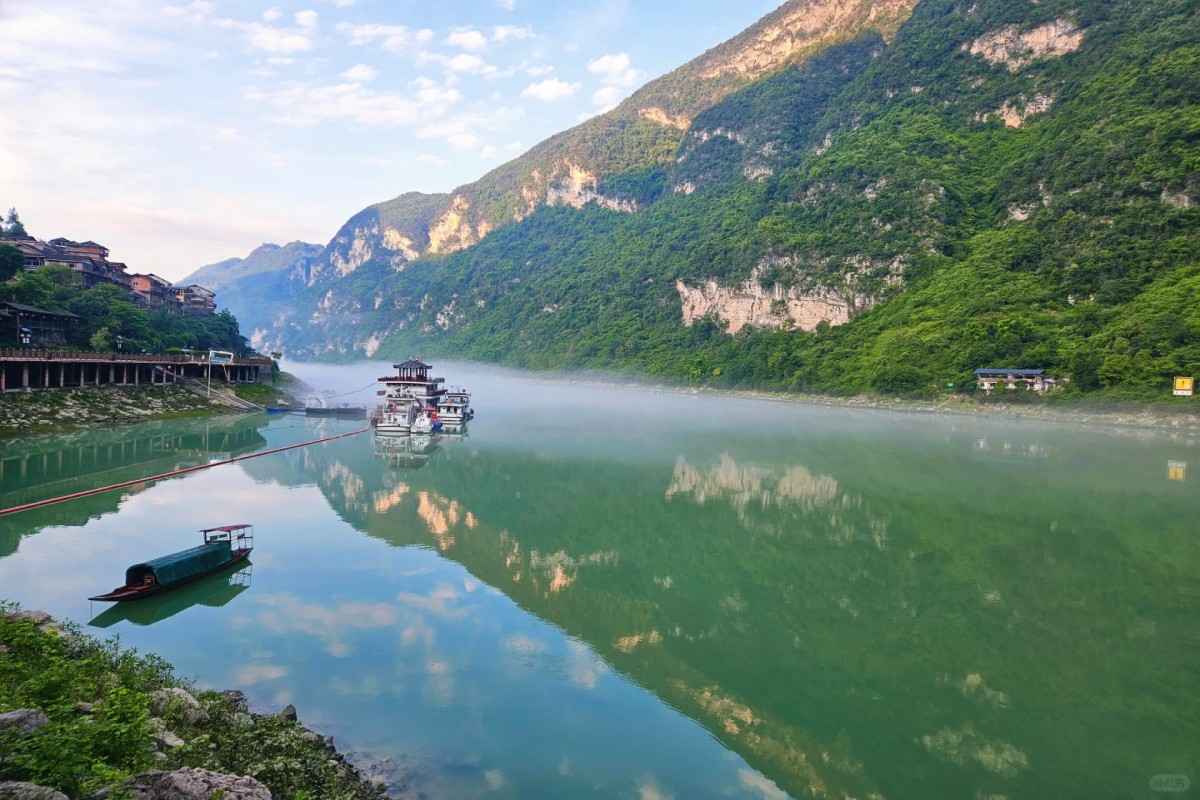 Calm green river with small boats and larger passenger vessels, reflecting steep forested mountains and riverside buildings under a partly cloudy sky with morning mist.