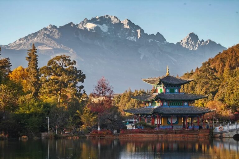 Traditional multi-tiered temple by a lake with autumn trees, reflecting on the water, with snow-capped mountains in the background.