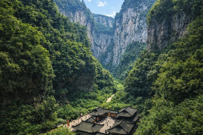 Traditional temple buildings at the base of a lush, steep mountain valley with cliffs and dense greenery, small crowds visible.