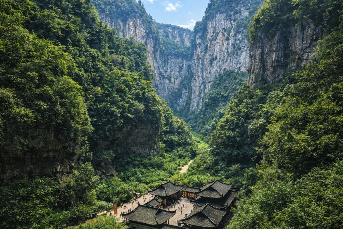 Traditional temple buildings at the base of a lush, steep mountain valley with cliffs and dense greenery, small crowds visible.