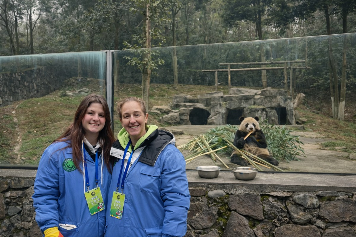 Two visitors posing in front of a giant panda eating bamboo behind glass at a panda conservation center.