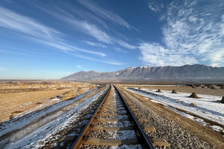 Railway track leading toward snowy mountains under a wide blue sky.
