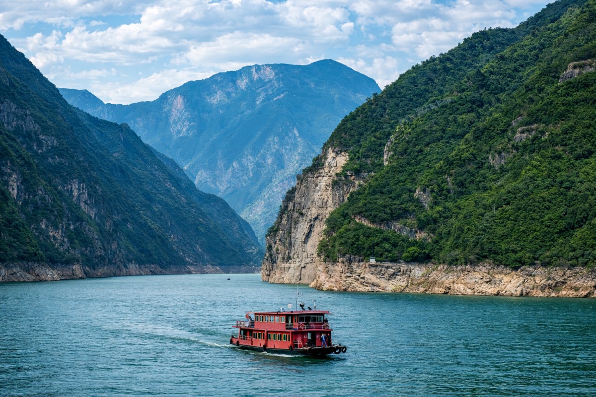Small red tourist boat cruising on river flanked by steep green cliffs under a partly cloudy sky.