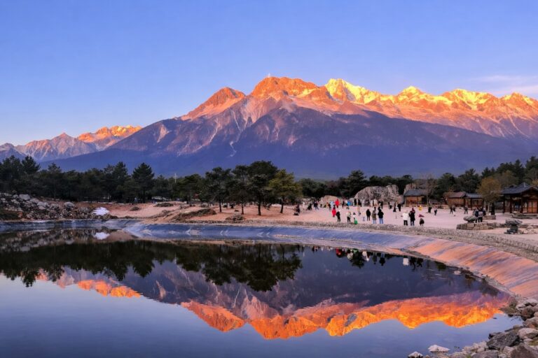 Tourists walking near a partially frozen lake with pine trees, reflecting snow-capped mountains glowing orange at sunrise.