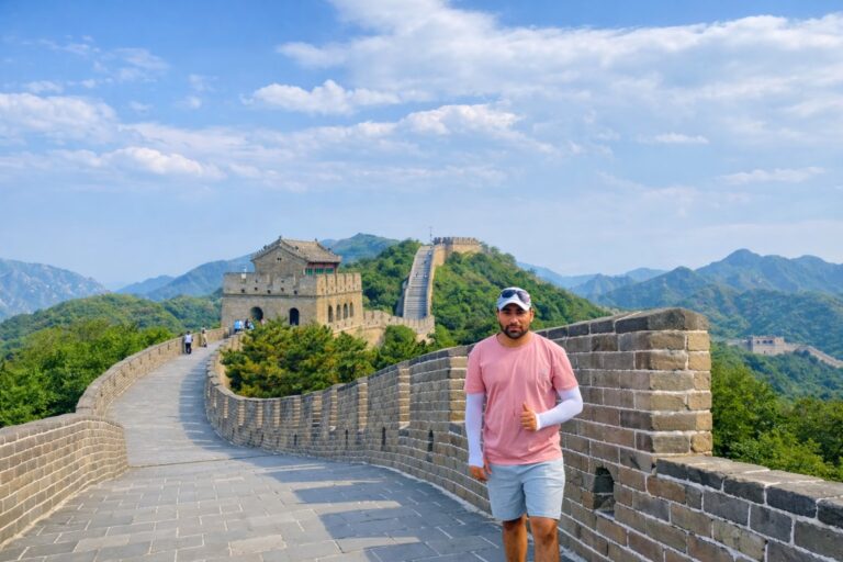 Seorang pria berbaju merah muda dan celana pendek mengacungkan jempol sambil berdiri di Tembok Besar China di bawah langit yang sebagian berawan.