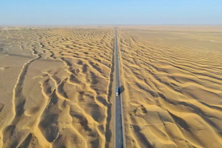 Long straight road cutting through vast sand dunes from above.