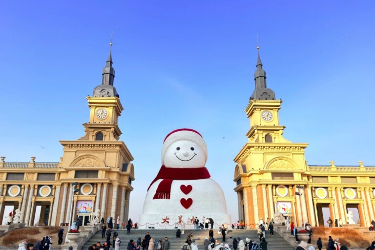 Large smiling snowman with red scarf in front of twin clock towers and crowds around.