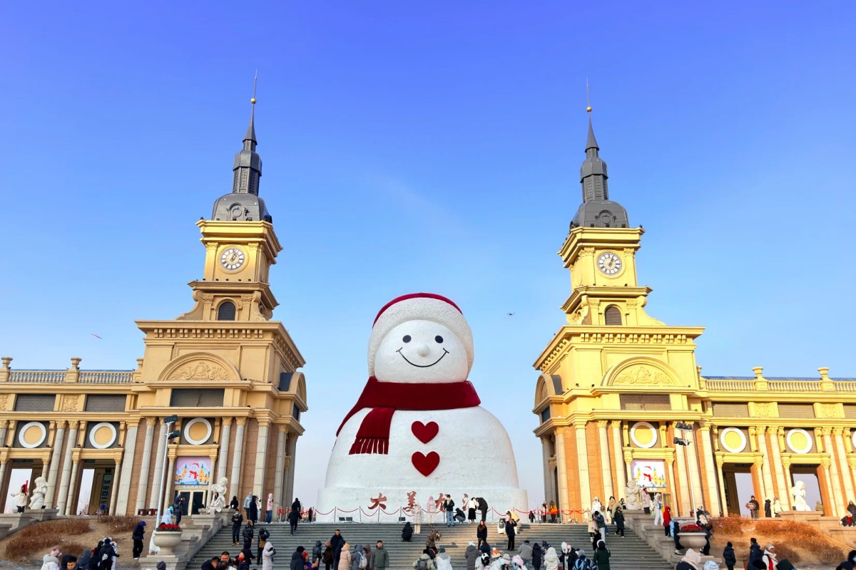 Large smiling snowman with red scarf in front of twin clock towers and crowds around.