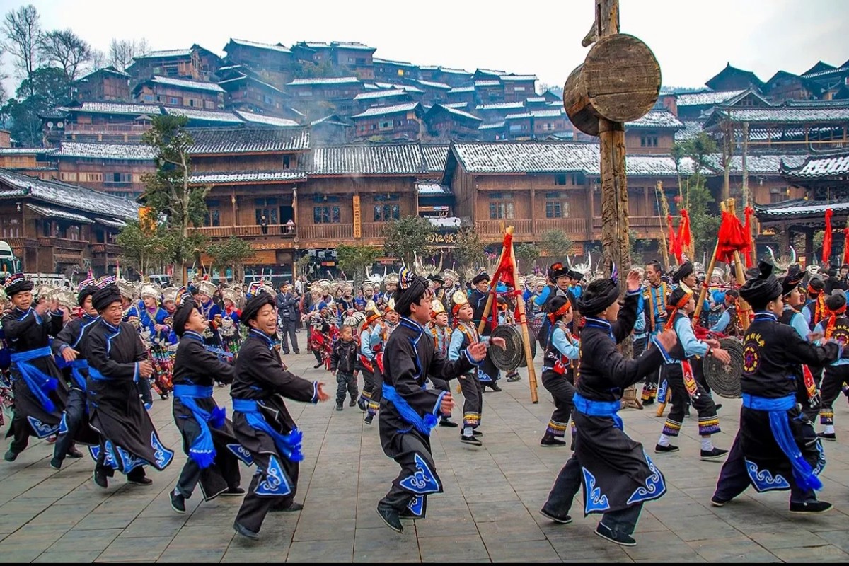 Miao men and women in traditional attire performing a drum and gong dance in a village square, with wooden houses on a hillside in the background.