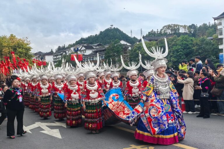 Miao women in traditional silver headdresses and colorful attire marching in a street parade, with spectators on both sides.