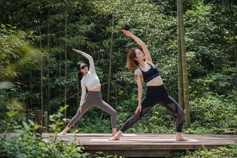 Two women doing yoga on a wooden deck in a green forest setting