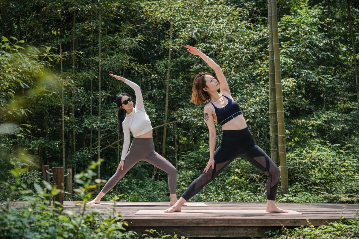 Two women doing yoga on a wooden deck in a green forest setting