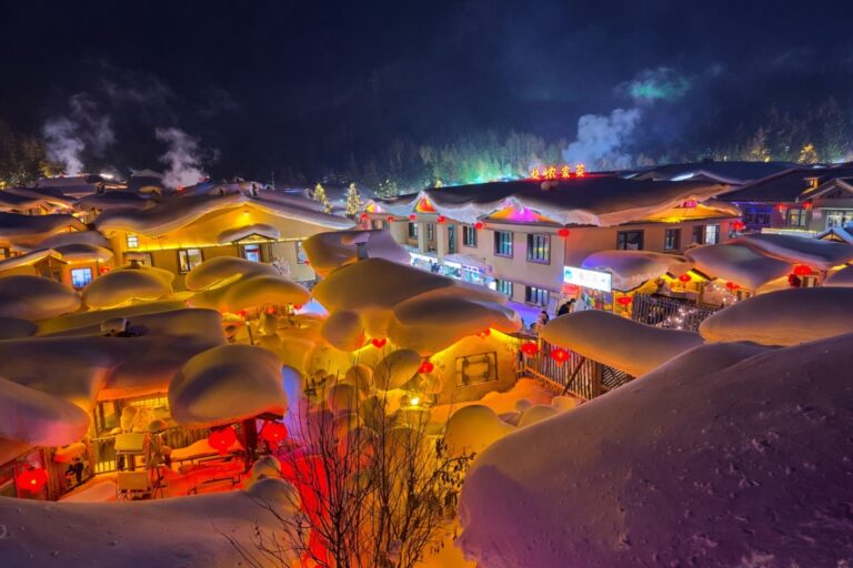 Snow-covered village at night with warm lights glowing from rooftops.