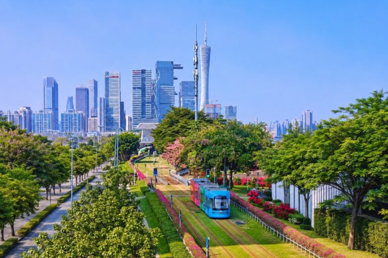 A modern tram travels through a green boulevard with the Guangzhou skyline and Canton Tower in the background.