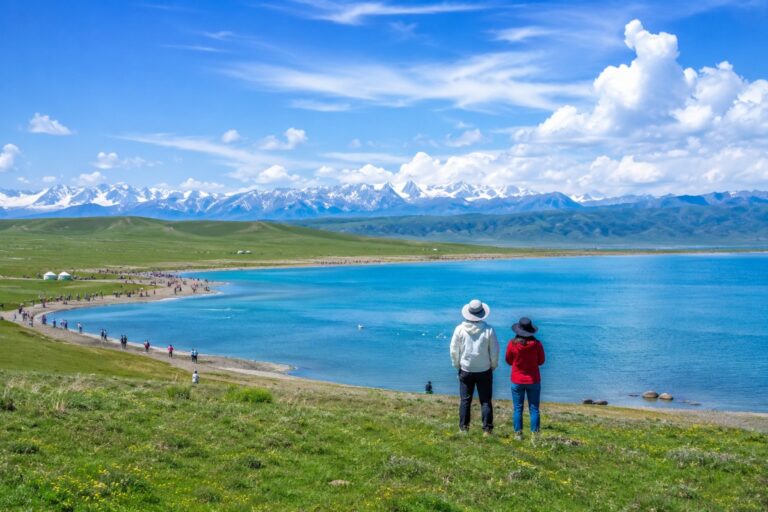 Travelers overlooking a blue alpine lake with green grasslands and snow-capped mountains under a clear sky.