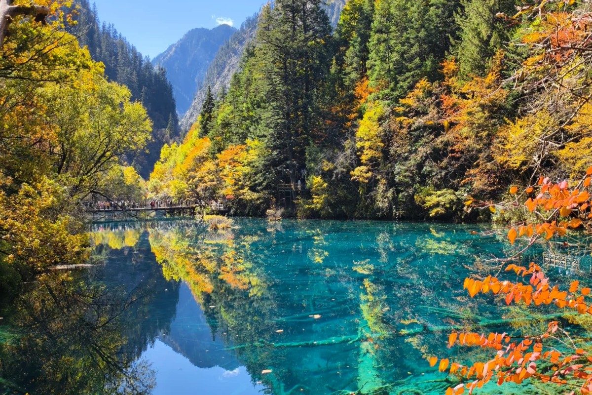 Crystal-clear turquoise lake surrounded by dense evergreen forest and vibrant autumn foliage, with colorful trees and mountain peaks reflected perfectly on the calm water under a bright blue sky.